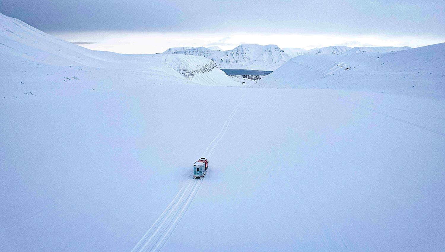 A snowcat on a glacier driving along tracks in the snow towards Longyearbyen in the background