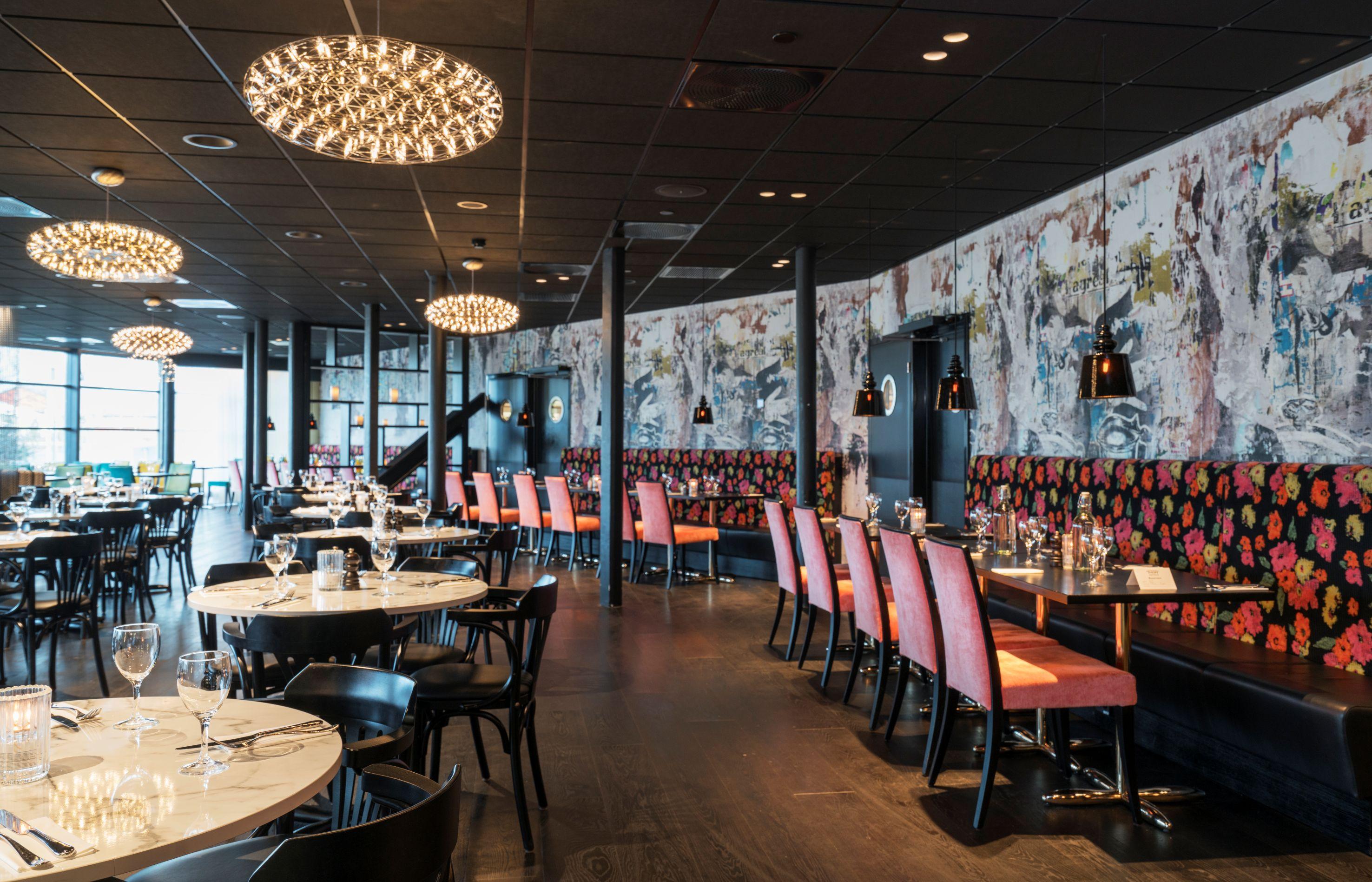 En moderne restaurant med runde og avlange bord, dekorative lamper og fargerike sitteplasser.