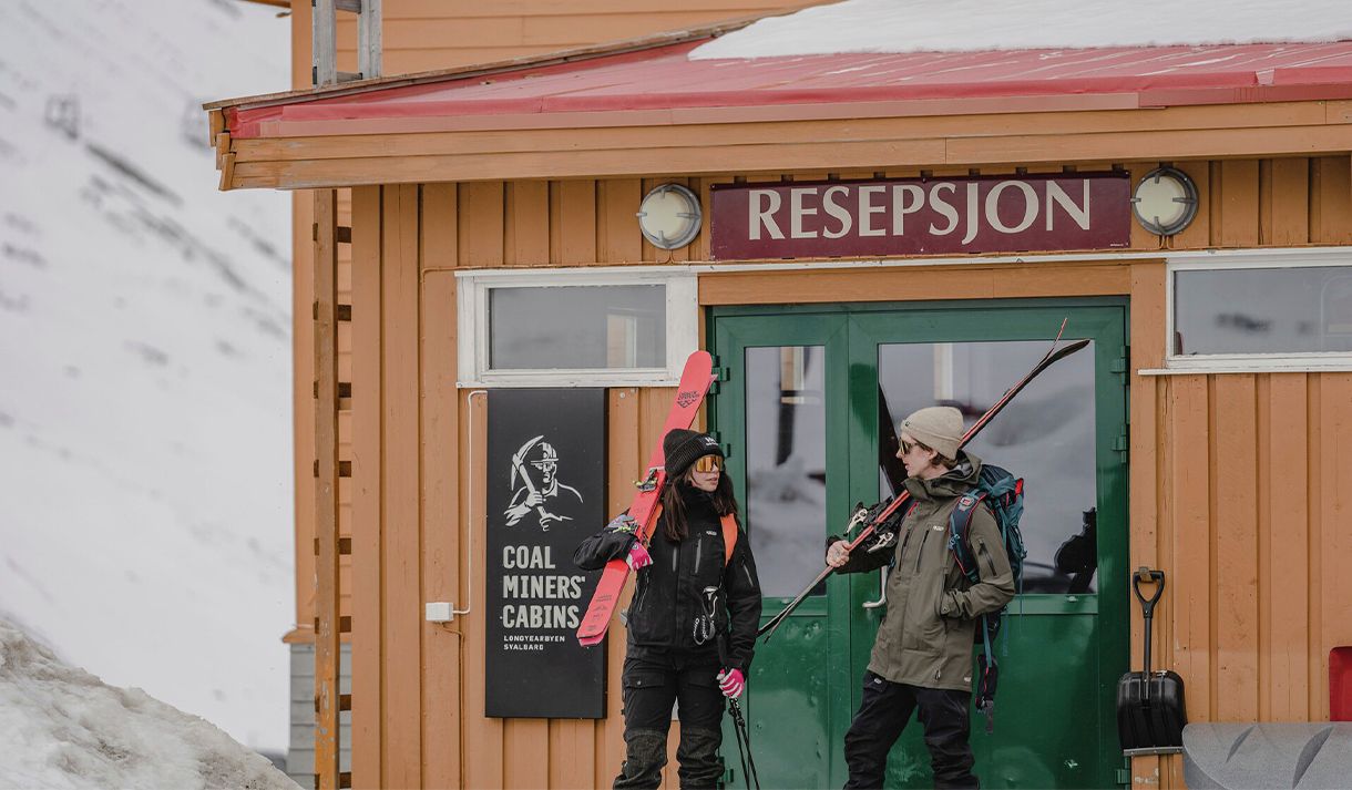 Two guests dressed for skiing carrying ski touring equipment outside the front door to the reception at Coal Miners' Cabins