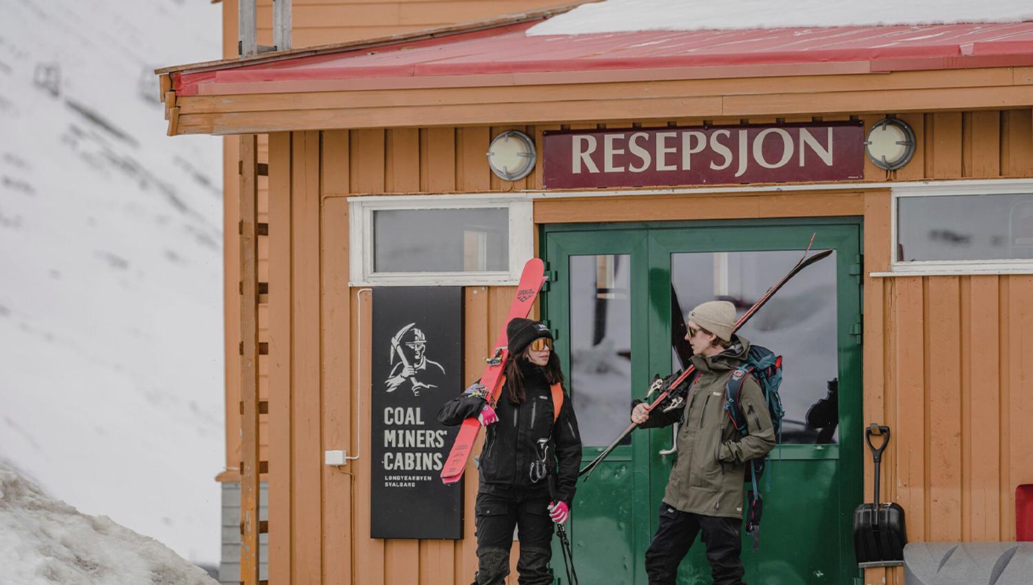 Two guests dressed for skiing carrying ski touring equipment outside the front door to the reception at Coal Miners' Cabins
