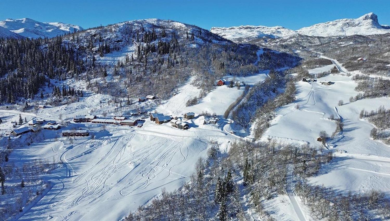 Luftbilde av en fjellstue i en solrik snødekt li omgitt av noe skog og med høye fjell i bakgrunnen