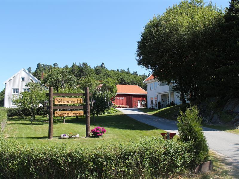 Innkjørsel til Sandnescampingen. Stort treskilt som står "Velkmmen til Sandes Camping" i en hage.