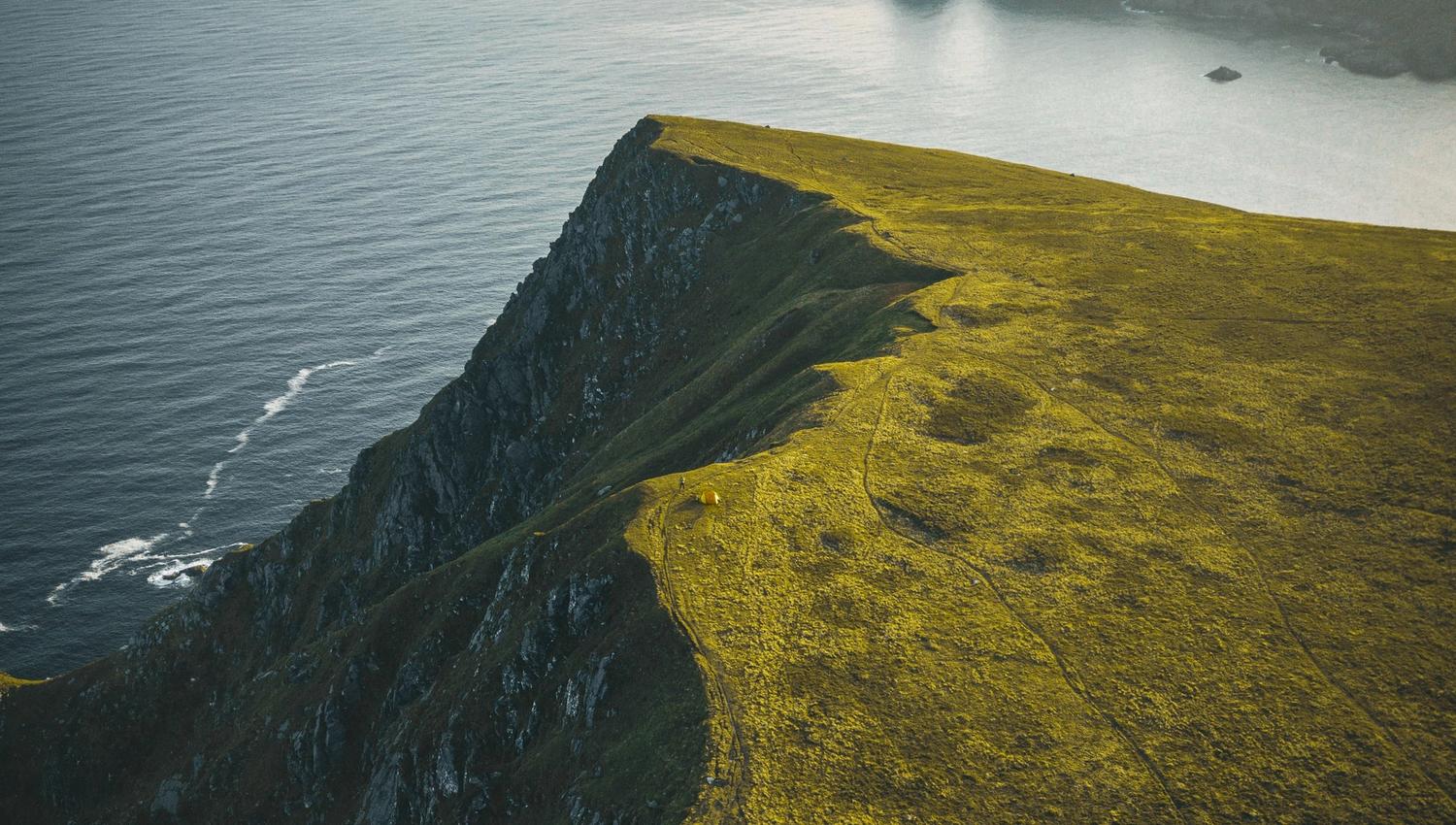 Solbelyst grasdekt klippe ved kysten med bratte stup ned mot havet, med øyar og dis i horisonten..