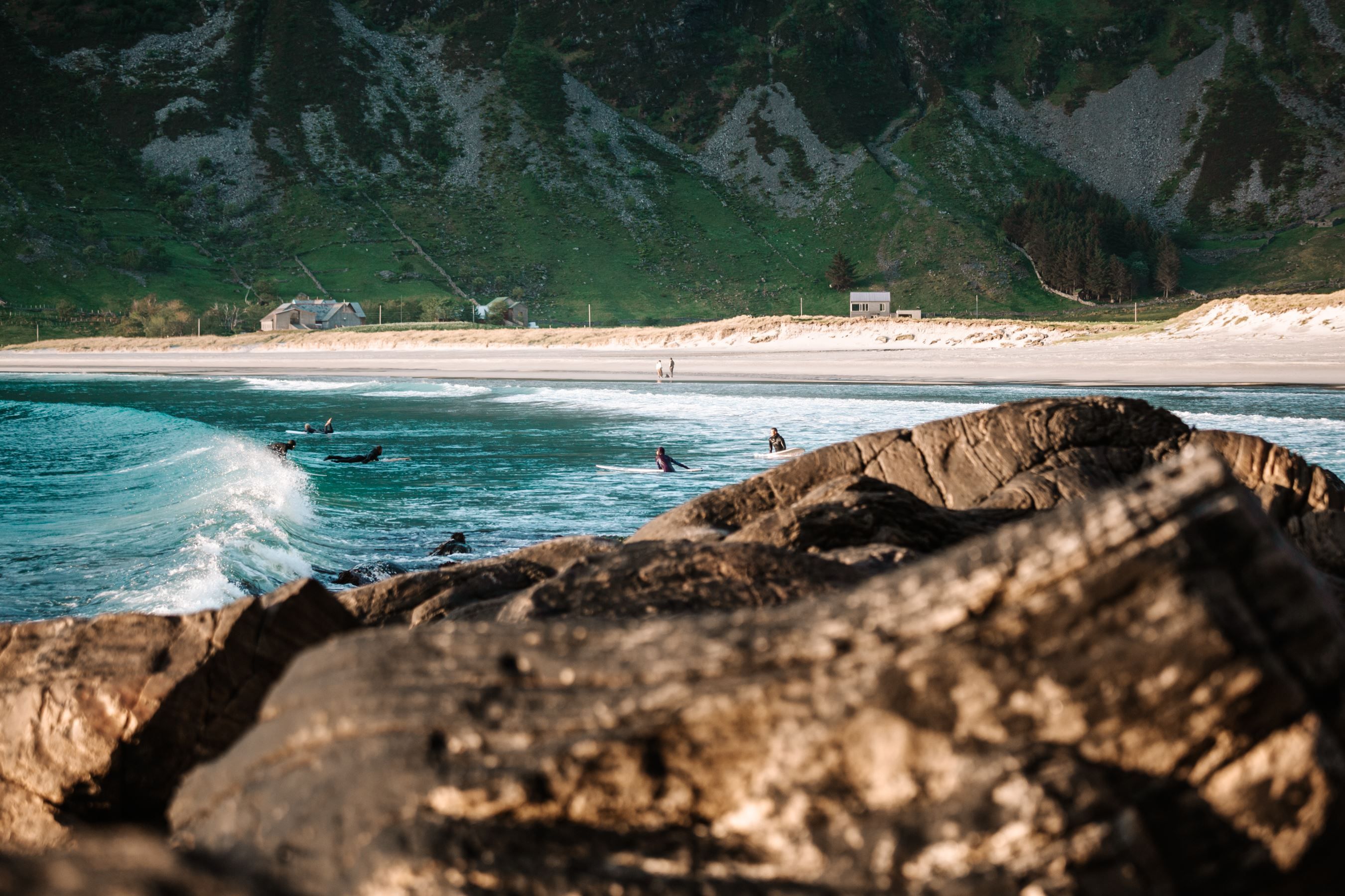 Surfers sit on boards in turquoise waves near a sandy beach, with rocky foreground and green mountains behind..