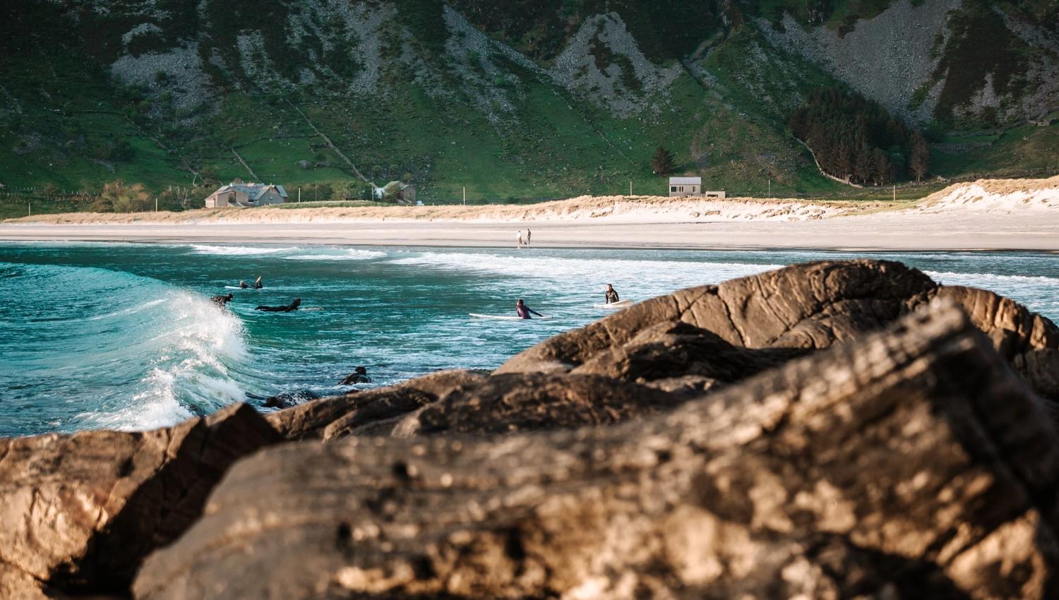 Surfers sit on boards in turquoise waves near a sandy beach, with rocky foreground and green mountains behind..