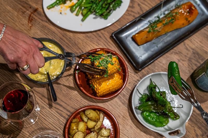 A hand using tongs over a table of small dishes like corn on the cob, potatoes, green peppers and sauce, with a glass of red wine nearby.