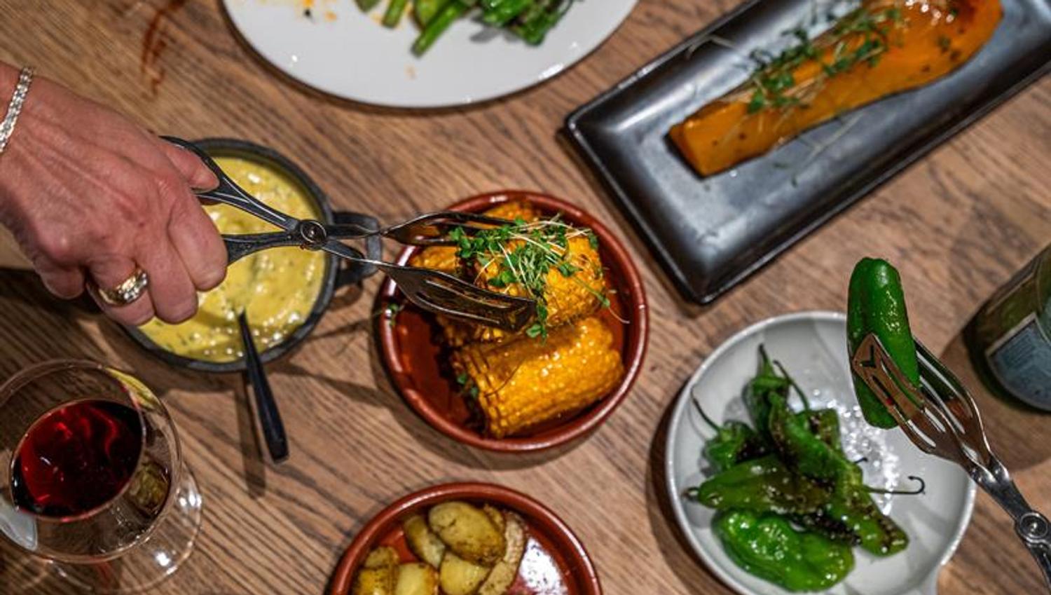 A hand using tongs over a table of small dishes like corn on the cob, potatoes, green peppers and sauce, with a glass of red wine nearby.