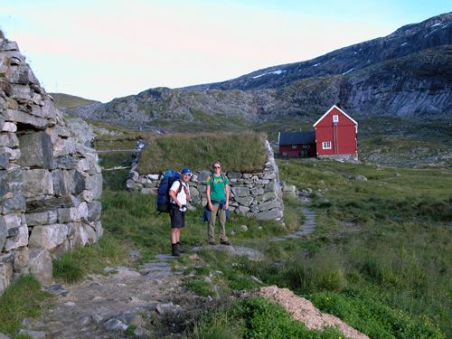 Hikers by a traditional red mountain lodge and old stone walls on the Hardangervidda high plateau.