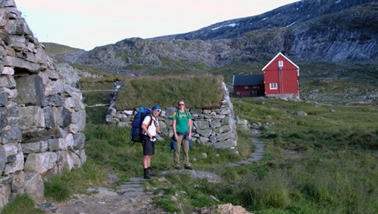 Hikers by a traditional red mountain lodge and old stone walls on the Hardangervidda high plateau.