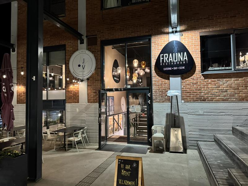 Inngangsparti til restauranten med uteservering, store vinduer og skiltet "Frauna". Stemningsfulle lys og dekor skaper en innbydende atmosfære.