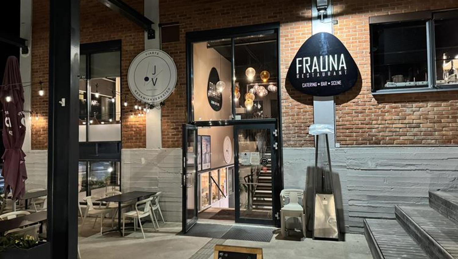 Inngangsparti til restauranten med uteservering, store vinduer og skiltet "Frauna". Stemningsfulle lys og dekor skaper en innbydende atmosfære.