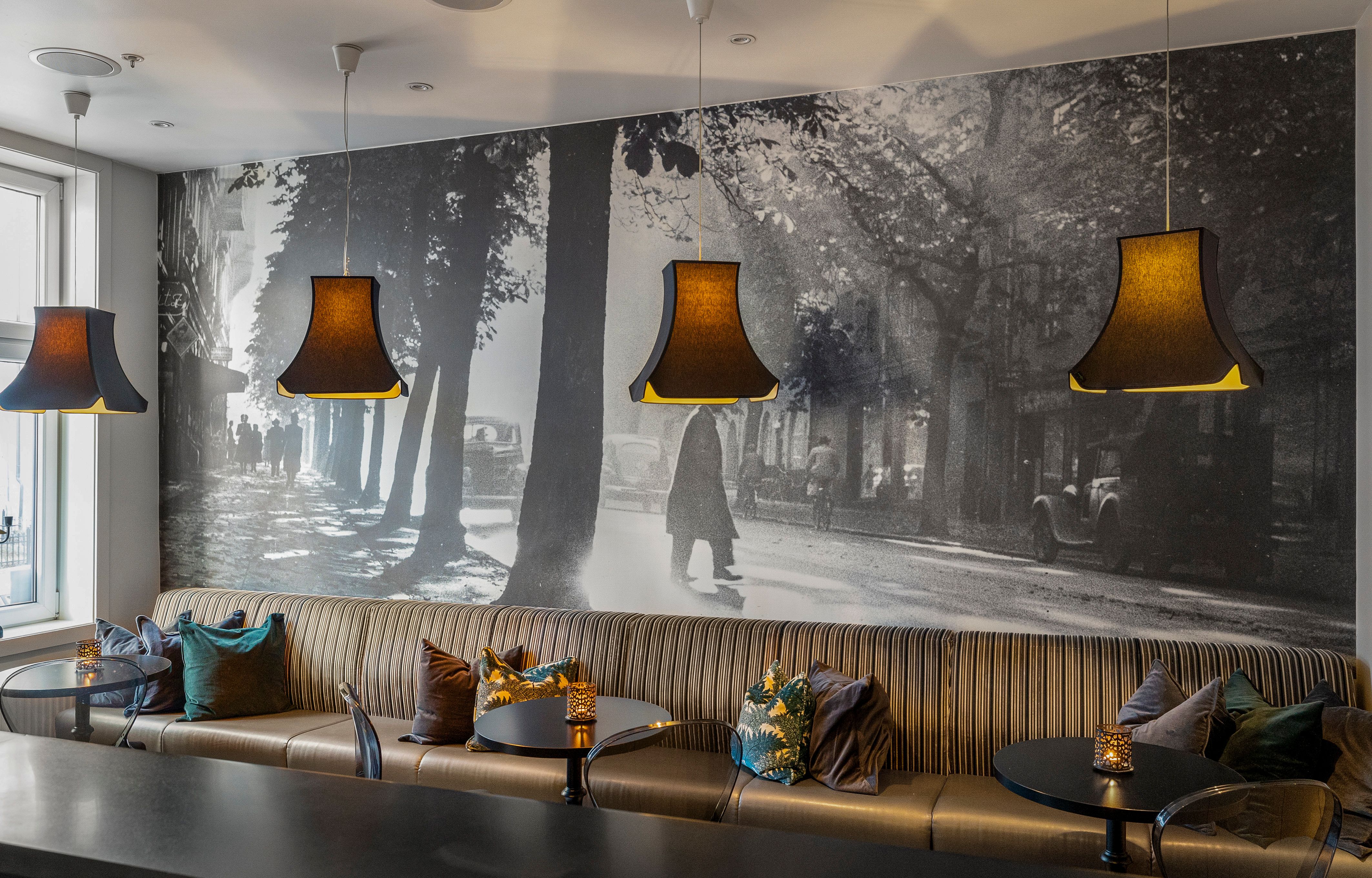 langsofa med puter. Fototapet med bymotiv. Brune lampetter over bordet. Saga Hotel Oslo. Foto