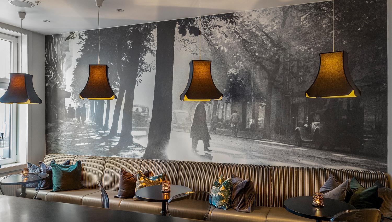 langsofa med puter. Fototapet med bymotiv. Brune lampetter over bordet. Saga Hotel Oslo. Foto