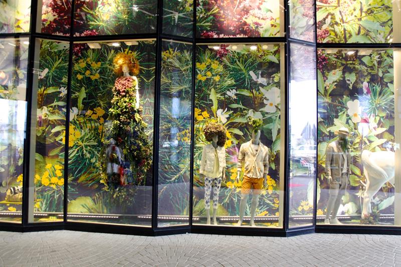 Butikkvindu med utstillingsdukker med klær foran en skogsbakgrunn.