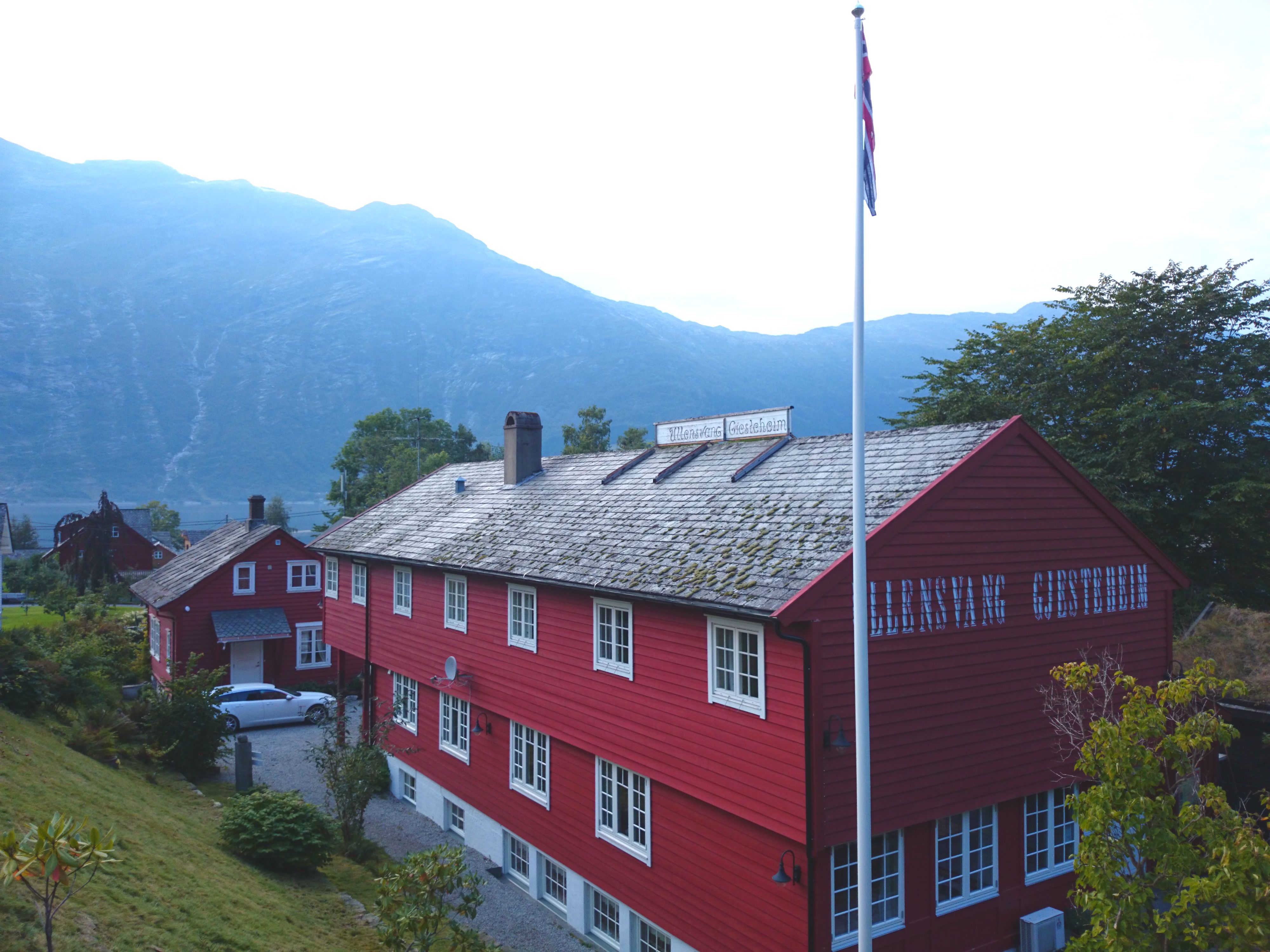 Ullensvang Gjesteheim sett frå tunet, med raudt trebygg, norsk flagg og fjellutsikt i bakgrunnen.