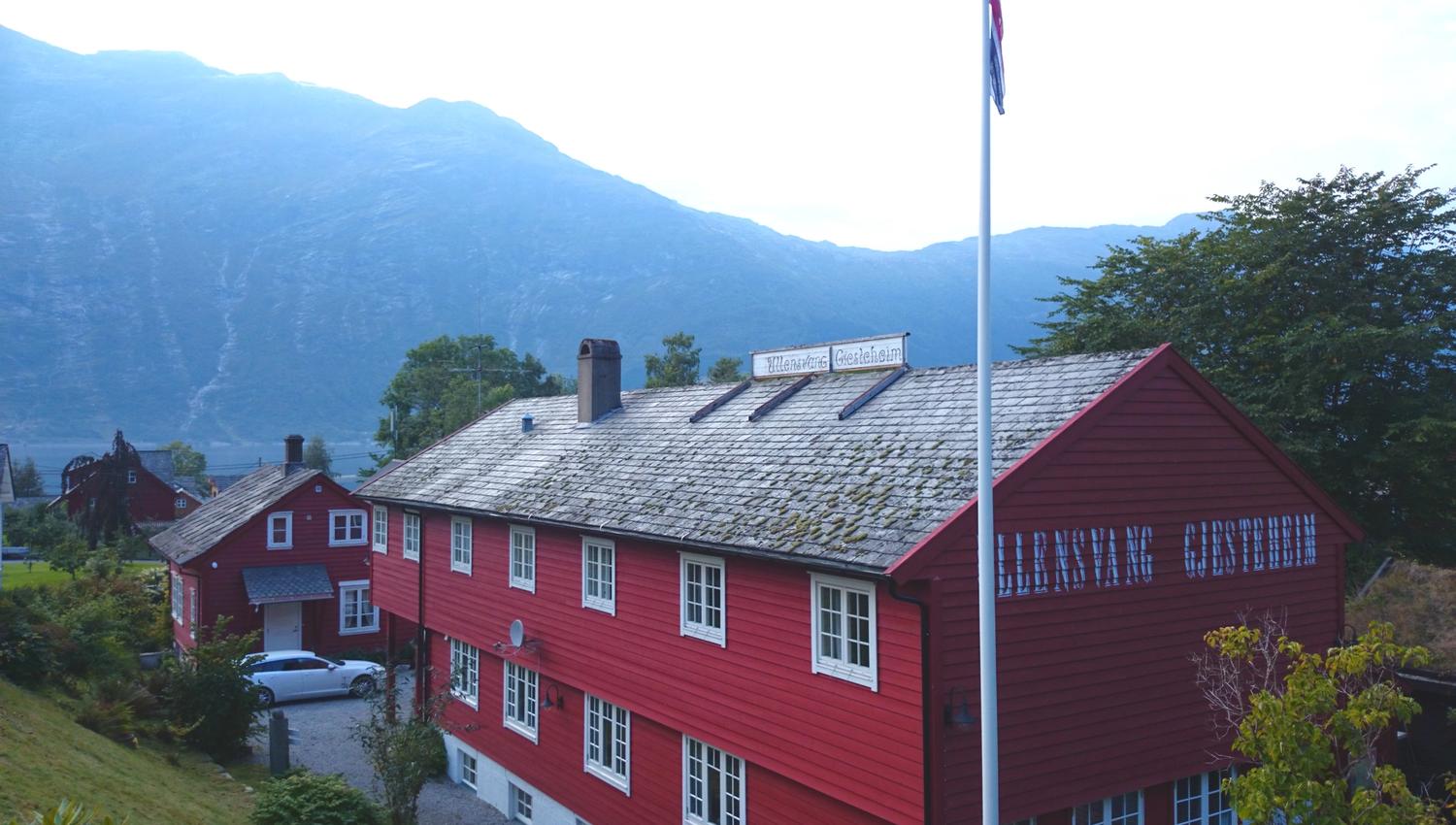 Ullensvang Gjesteheim sett frå tunet, med raudt trebygg, norsk flagg og fjellutsikt i bakgrunnen.