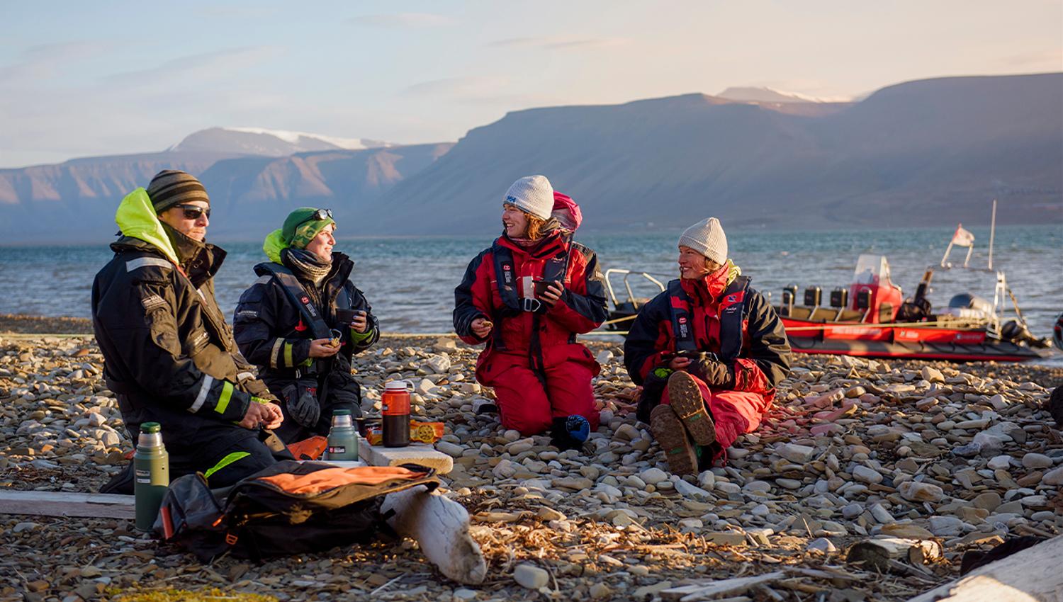 Two guides and two guests sitting on a beach talking with each other, and a RIB boat anchored by the shore of the fjord in the background