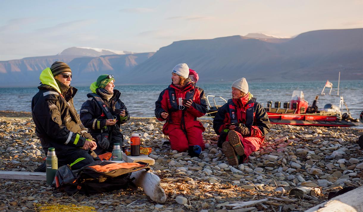 To guider og to gjester som sitter på en strand og prater med en RIB båt ankret langs sjøkanten i fjorden i bakgrunnen