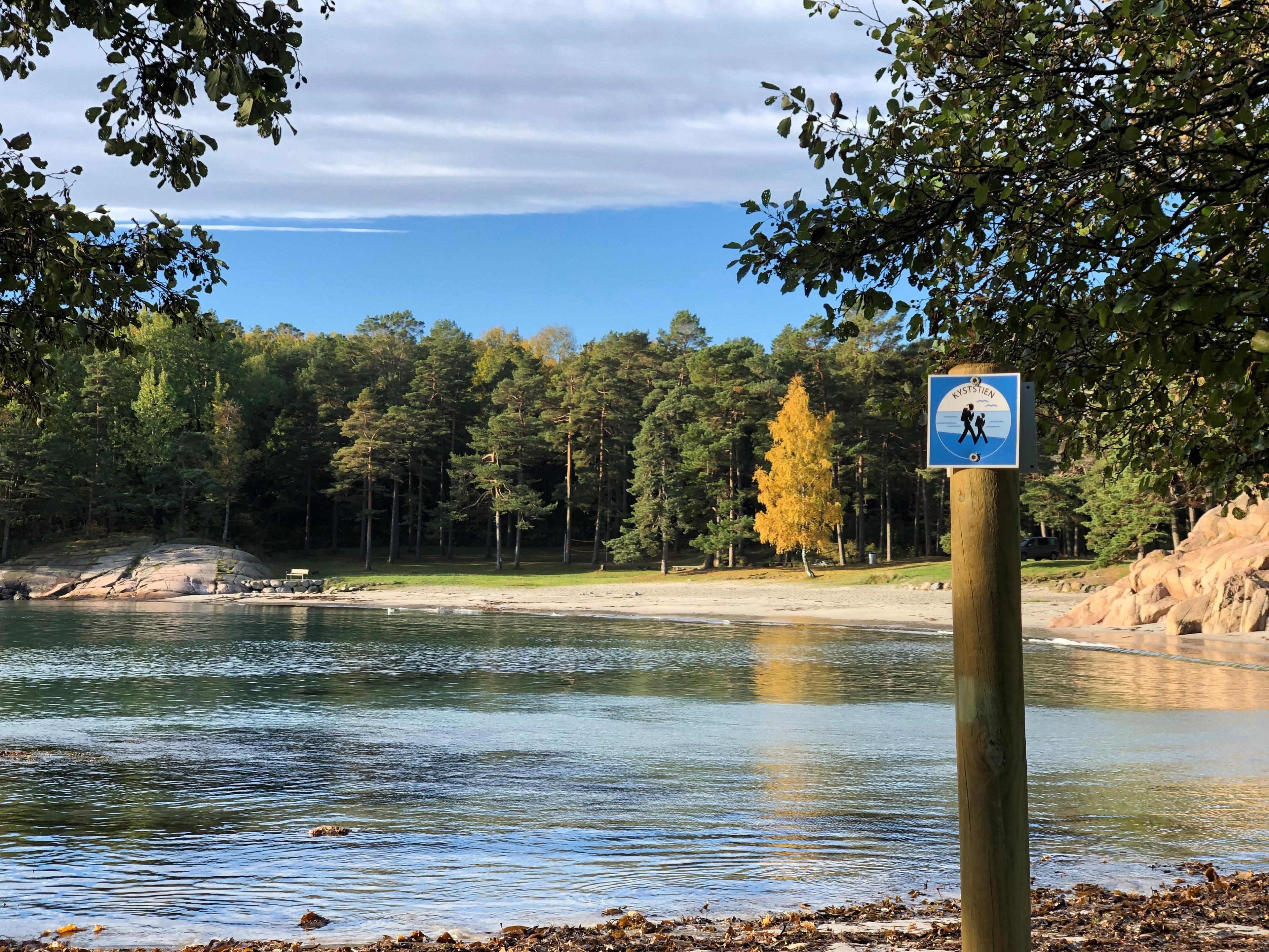 Bukt med strand og trær i bakgrunnen. Et liste skilt med "kyststien" fremme i bildet.