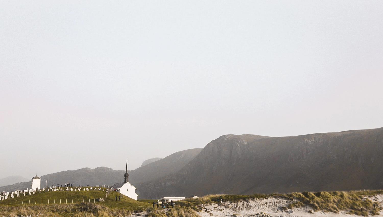 Kvit kyrkje og gravplass på ei grøn høgd ved ein steinete fjøre, med fjell i bakgrunnen og lys himmel..