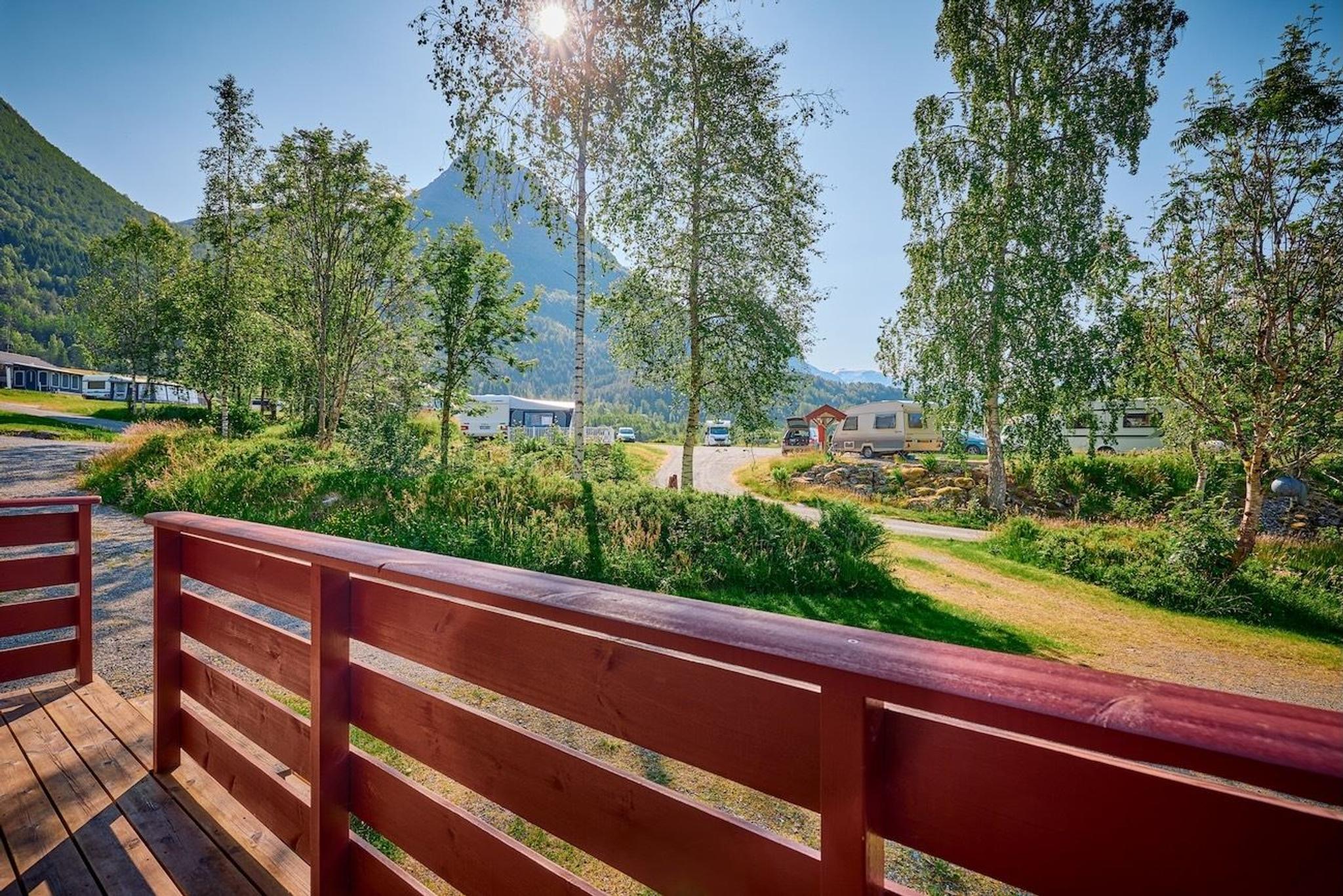 Red wooden deck railing overlooks a green campground with trees, caravans, and mountains under bright sun..
