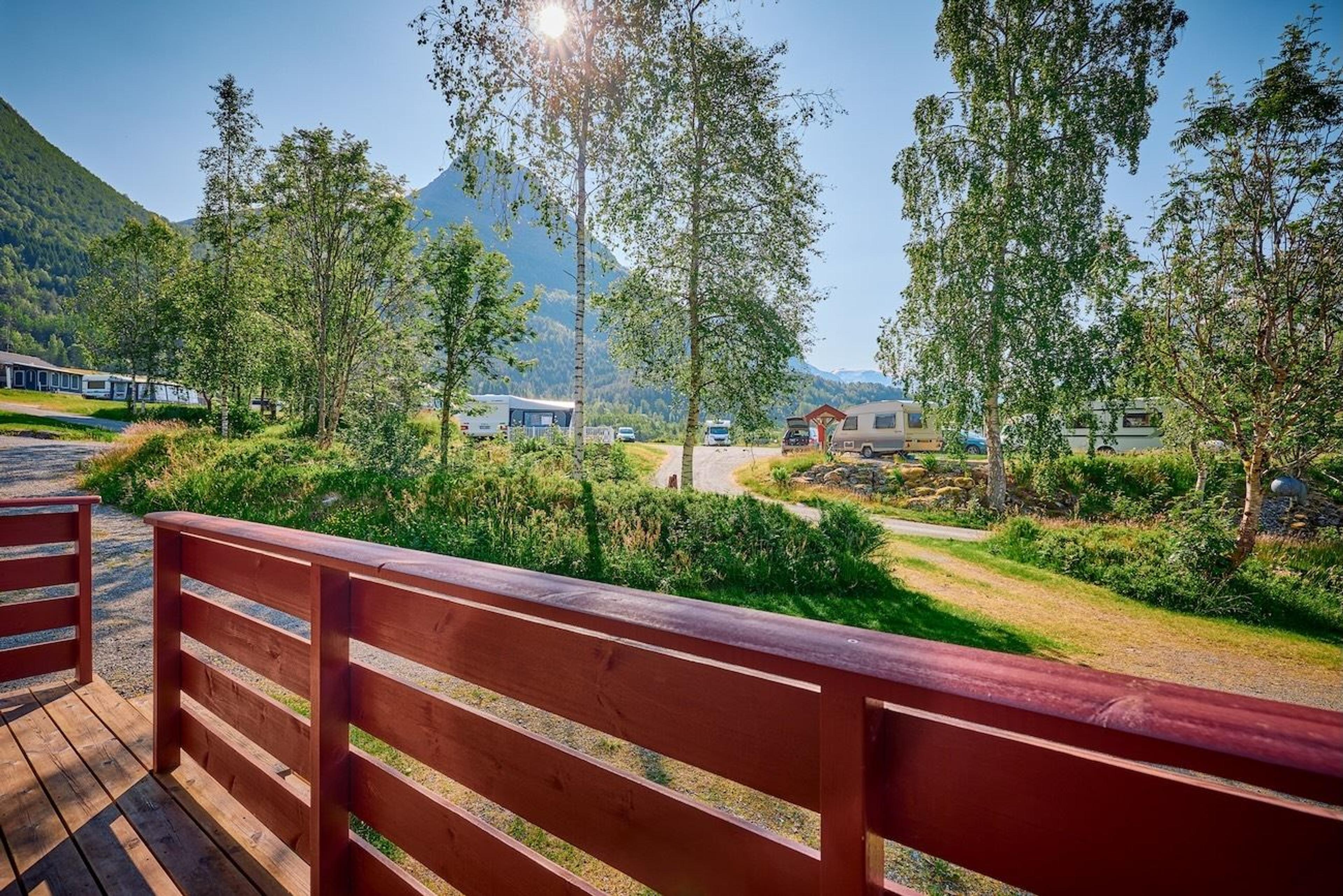 Red wooden deck railing overlooks a green campground with trees, caravans, and mountains under bright sun..