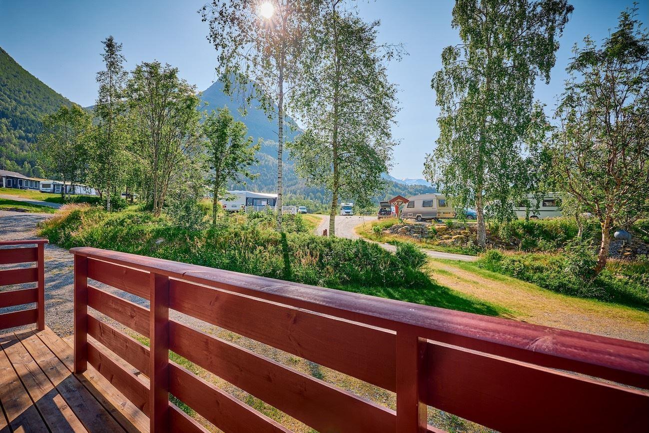 Red wooden deck railing overlooks a green campground with trees, caravans, and mountains under bright sun..