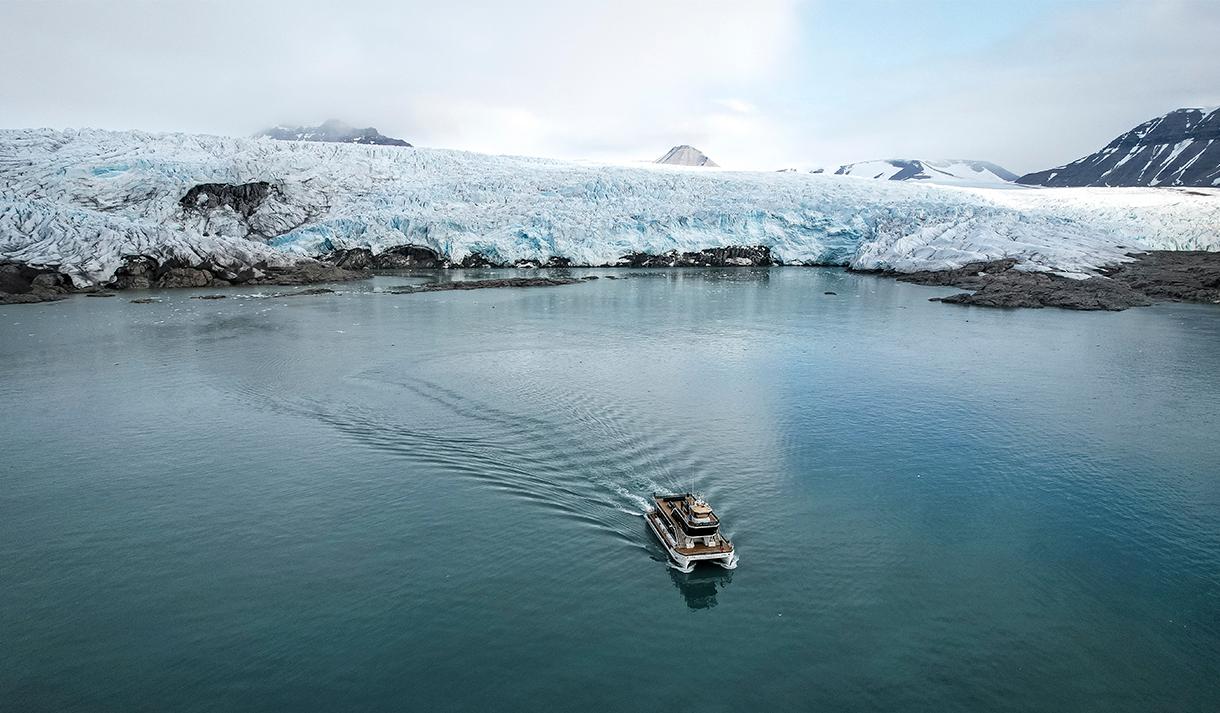 A boat sailing on a fjord in the foreground with a glacier front and mountains surrounding a bay in the background