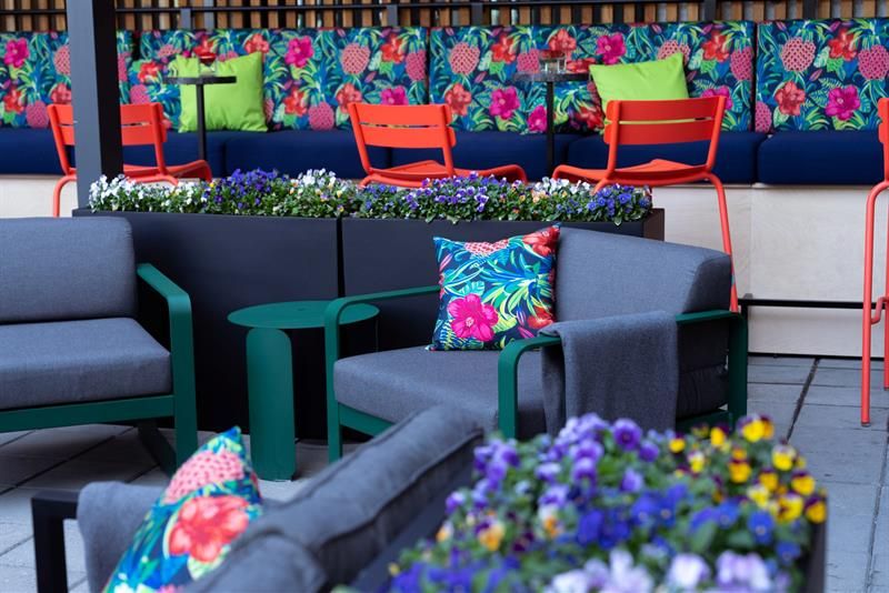 An outdoor seating area with grey-cushioned chairs, flowers, red chairs, and a floral seating nook in the background