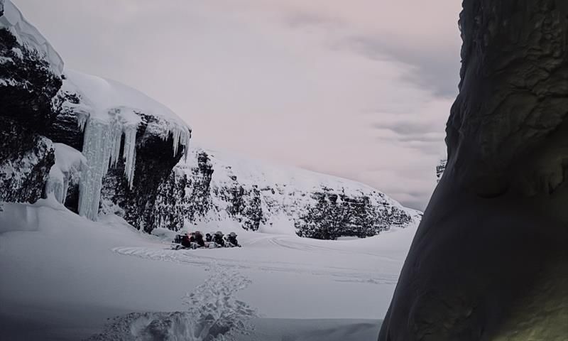 Picture taken from inside a cave, toward a opening with a  cliff on the left, snowmobiles standing on the flat beside it.
