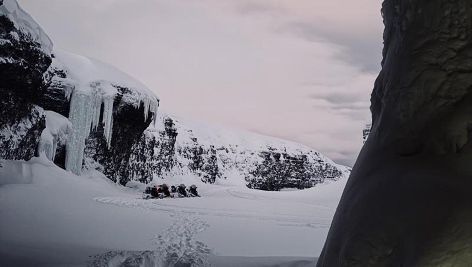 Picture taken from inside a cave, toward a opening with a cliff on the left, snowmobiles standing on the flat beside it.