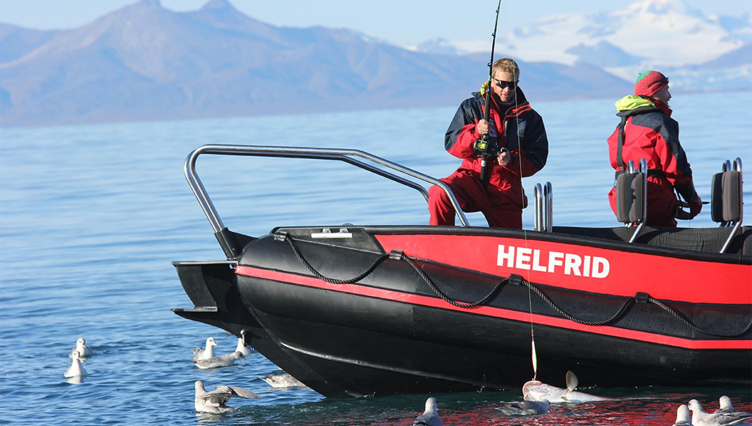 Two persons on board a RIB boat where one is holding a fishing pole with a fish on the hook floating on the water's surface in the foreground, surroun
