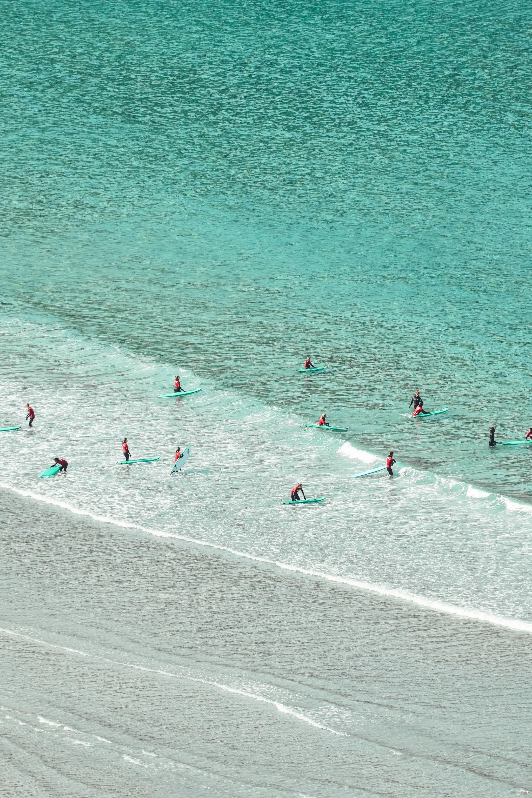 Several surfers in wetsuits sit on surfboards near shore as small waves roll over a sandy beach..