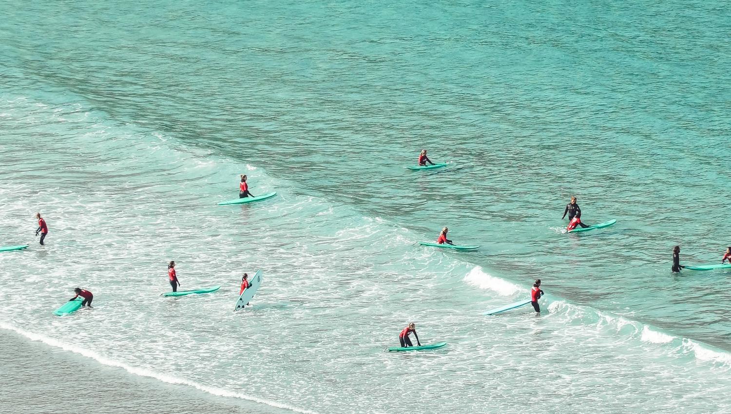 Several surfers in wetsuits sit on surfboards near shore as small waves roll over a sandy beach..