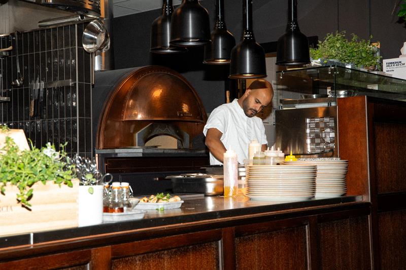 A chef prepares food at the counter in front of a wood-fired oven in a modern restaurant kitchen.