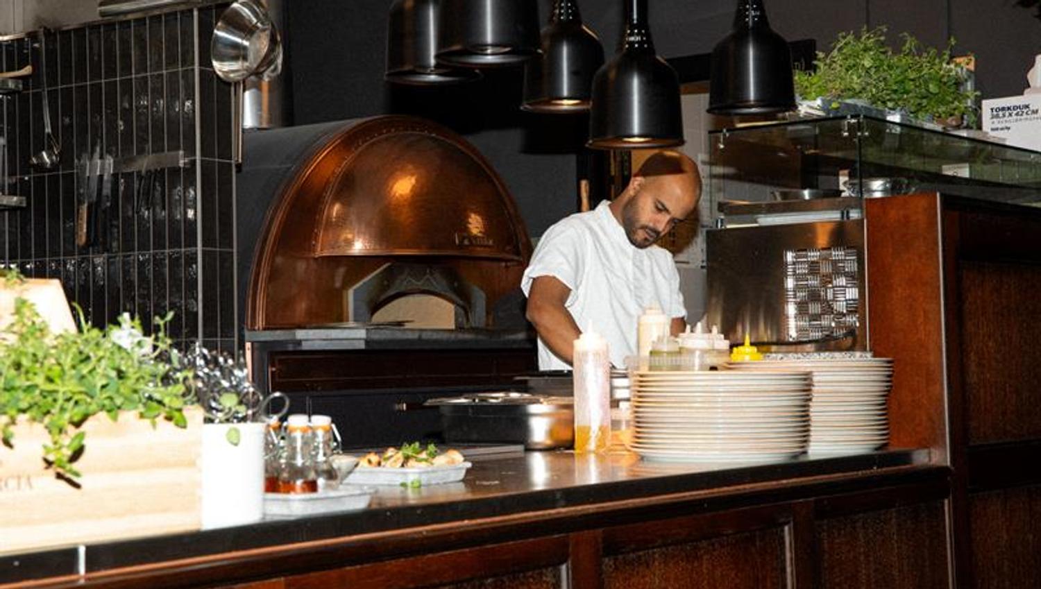 A chef prepares food at the counter in front of a wood-fired oven in a modern restaurant kitchen.