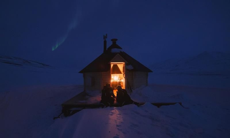 To personer sitter utenfor en koie med åpen dør. Inne brenner åpen ild i ovnen. Nordlys på himmelen.