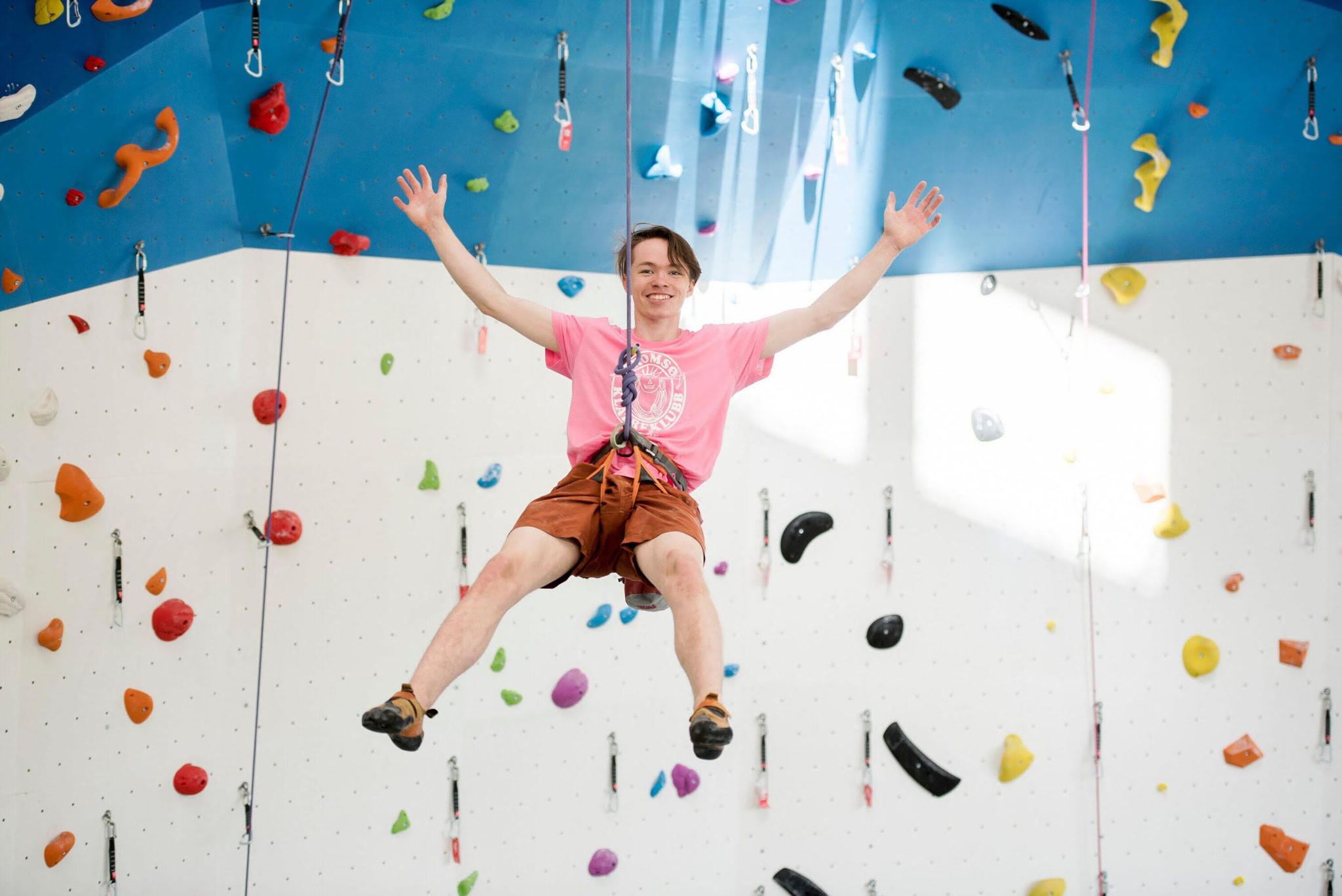 Person climbing an indoor climbing wall with colourful holds, wearing a pink shirt and orange shorts.