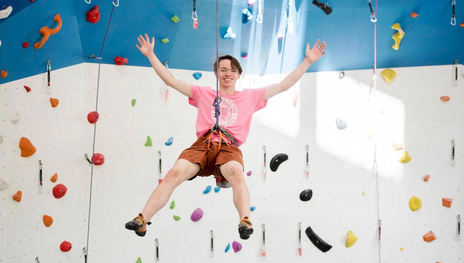 Person climbing an indoor climbing wall with colourful holds, wearing a pink shirt and orange shorts.