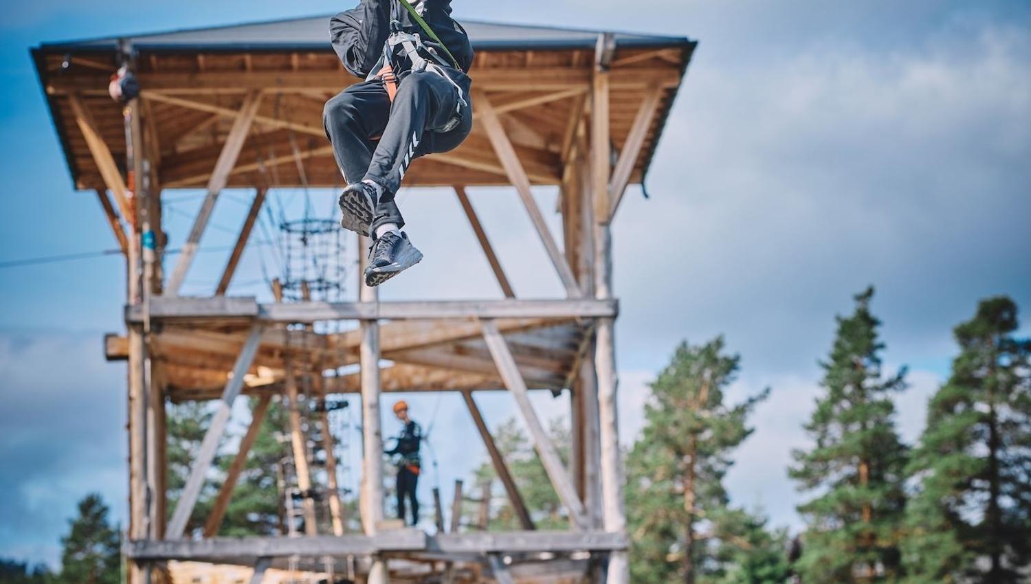 Ein person med hjelm og sele glir i zipline framfor eit høgt tretårn, med skog og blå himmel i bakgrunnen..
