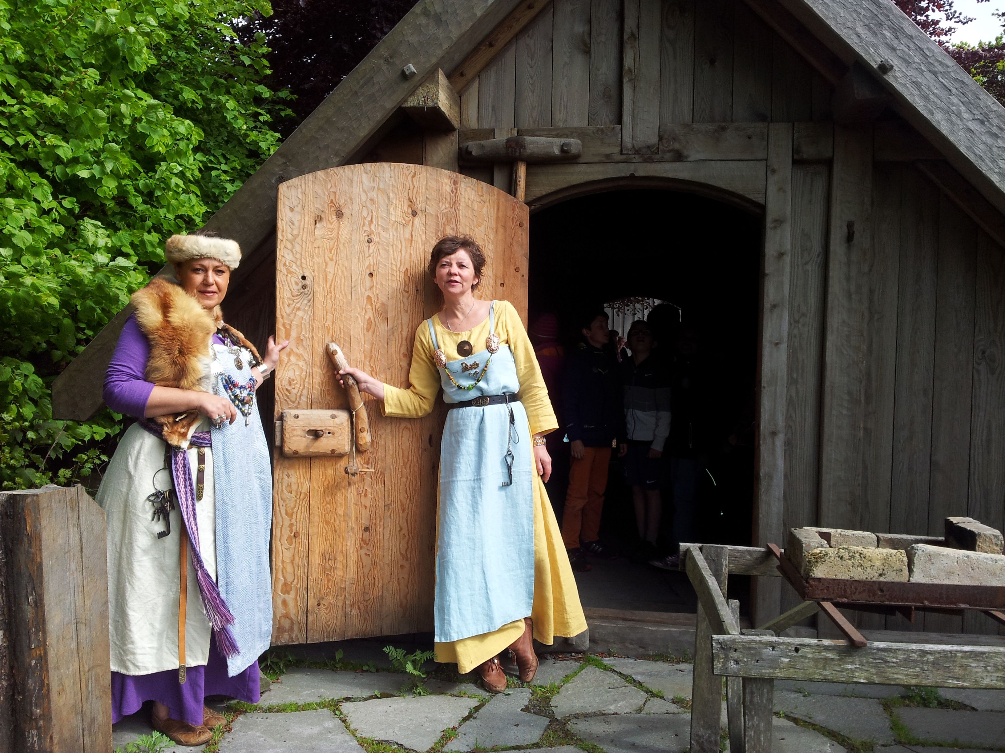 Two ladies dressed as Vikings stand in front of a Viking house and welcome you.