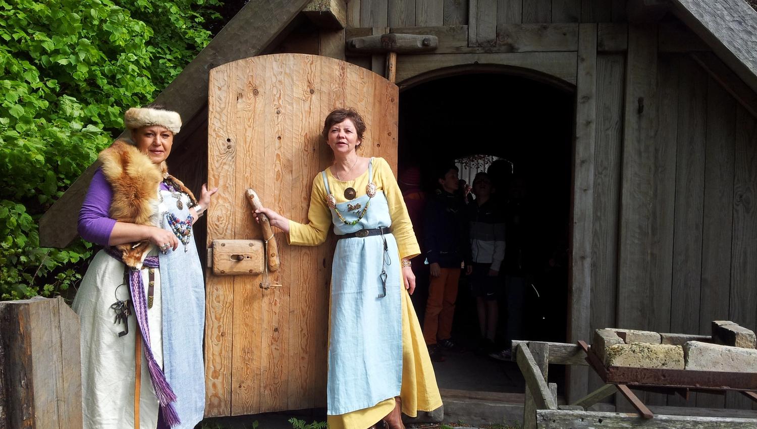Two ladies dressed as Vikings stand in front of a Viking house and welcome you.
