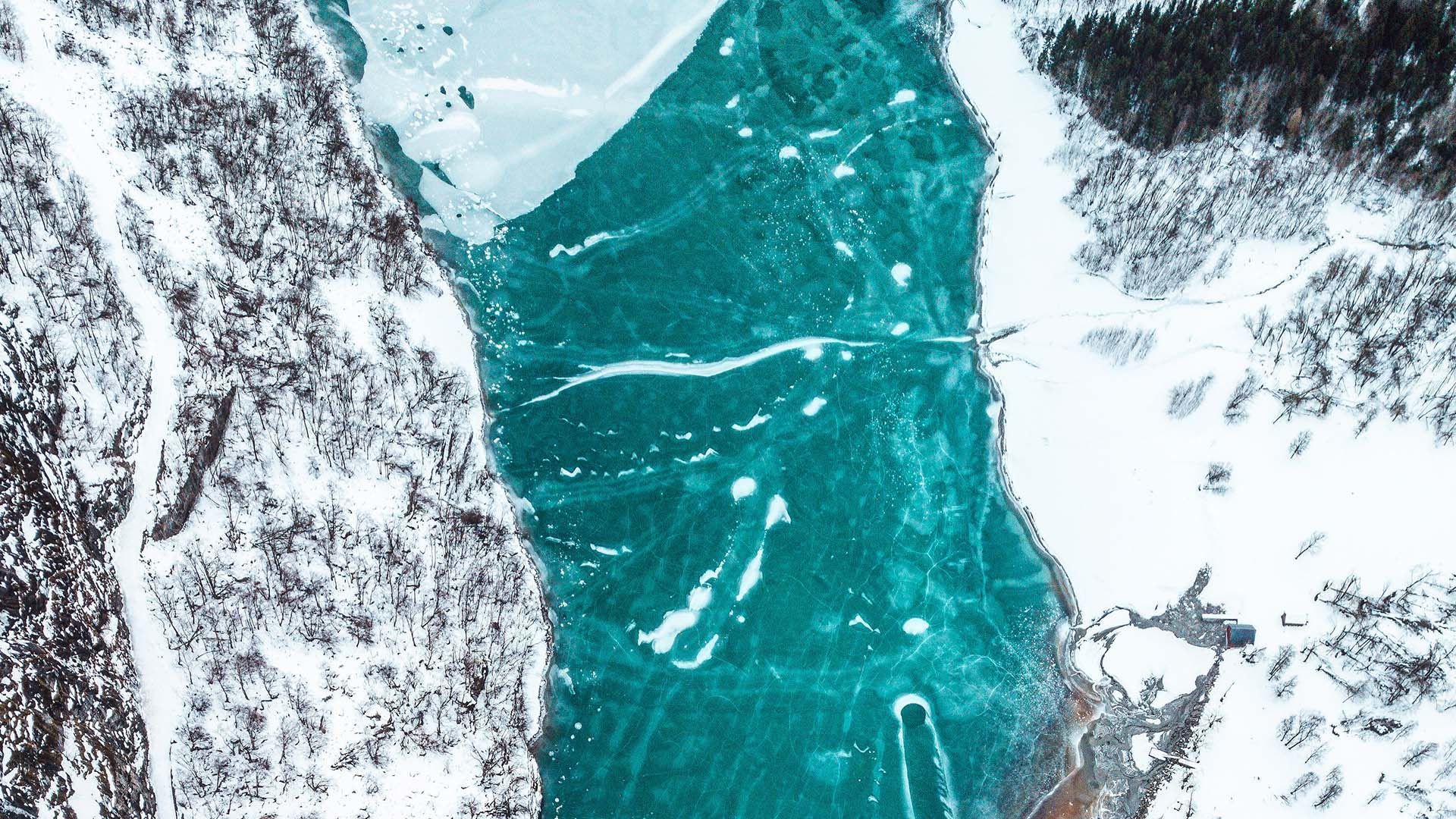 Luftfoto av turkis islagt elv mellom snødekte skogkledde skråningar, med sprekkar og isflak i overflata i Lodalen på Lovatnet