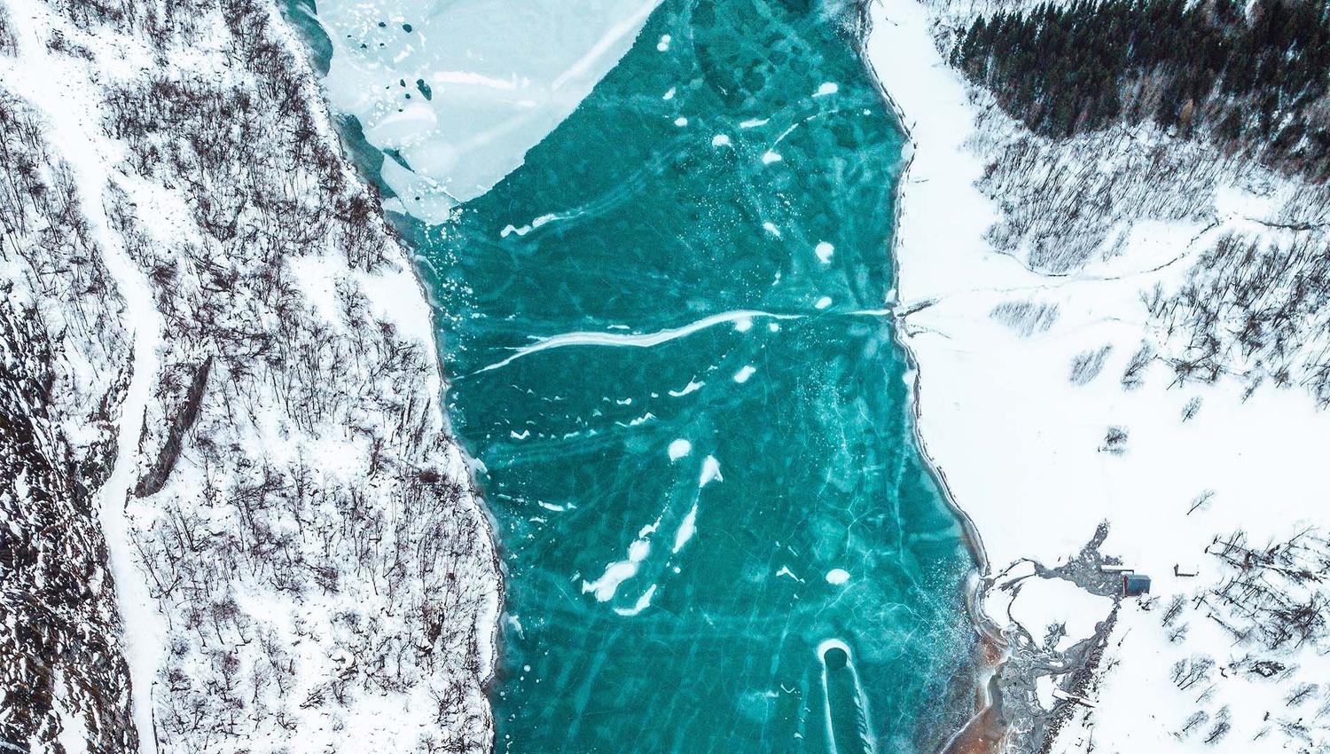 Luftfoto av turkis islagt elv mellom snødekte skogkledde skråningar, med sprekkar og isflak i overflata i Lodalen på Lovatnet