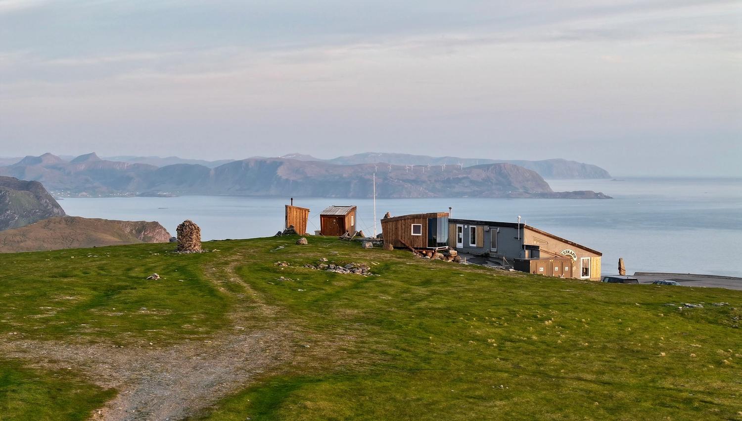 Grøn grasbakke med sti mot ei lita hytte og uthus ved fjord, med fjelløy og roleg hav i bakgrunnen..