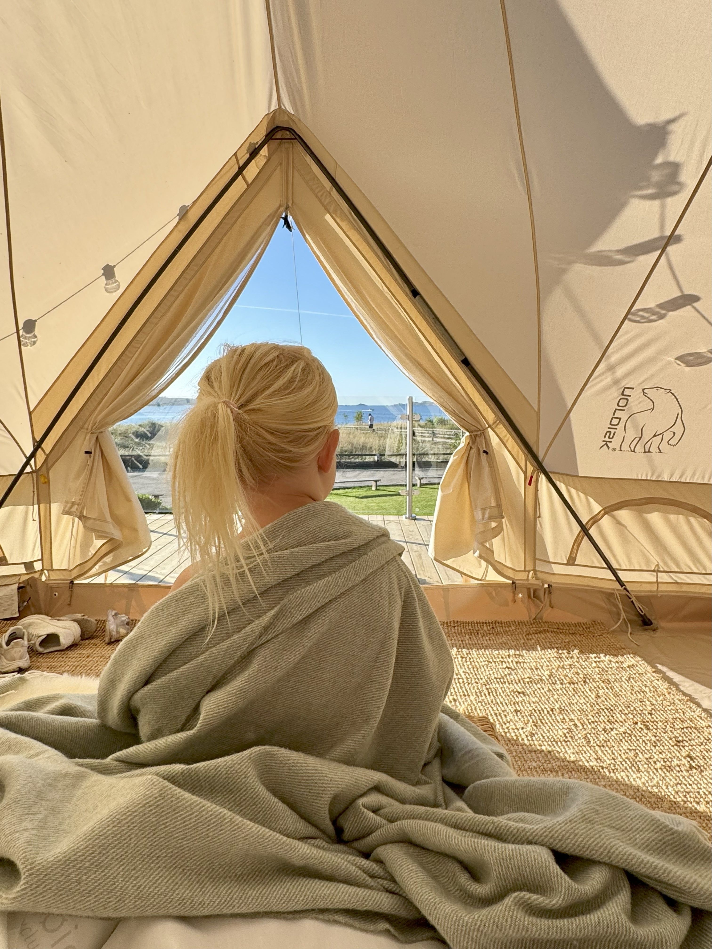 Liten jente som sitter i glampingteltet, innpakket i et teppe og kikker ut på utsikten ut mot havet