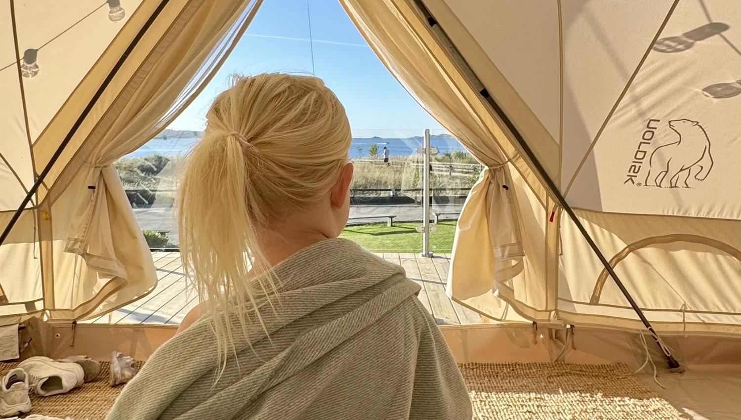 Liten jente som sitter i glampingteltet, innpakket i et teppe og kikker ut på utsikten ut mot havet