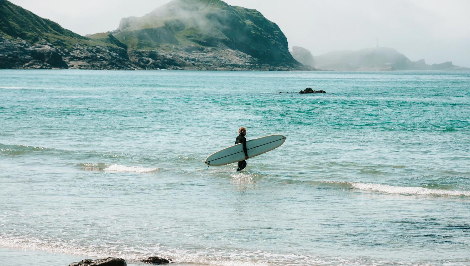 Ein surfear i Ervik i våtdrakt vassar i grunt hav med surfebrett, med klipper og tåkelagde fjell i bakgrunnen..