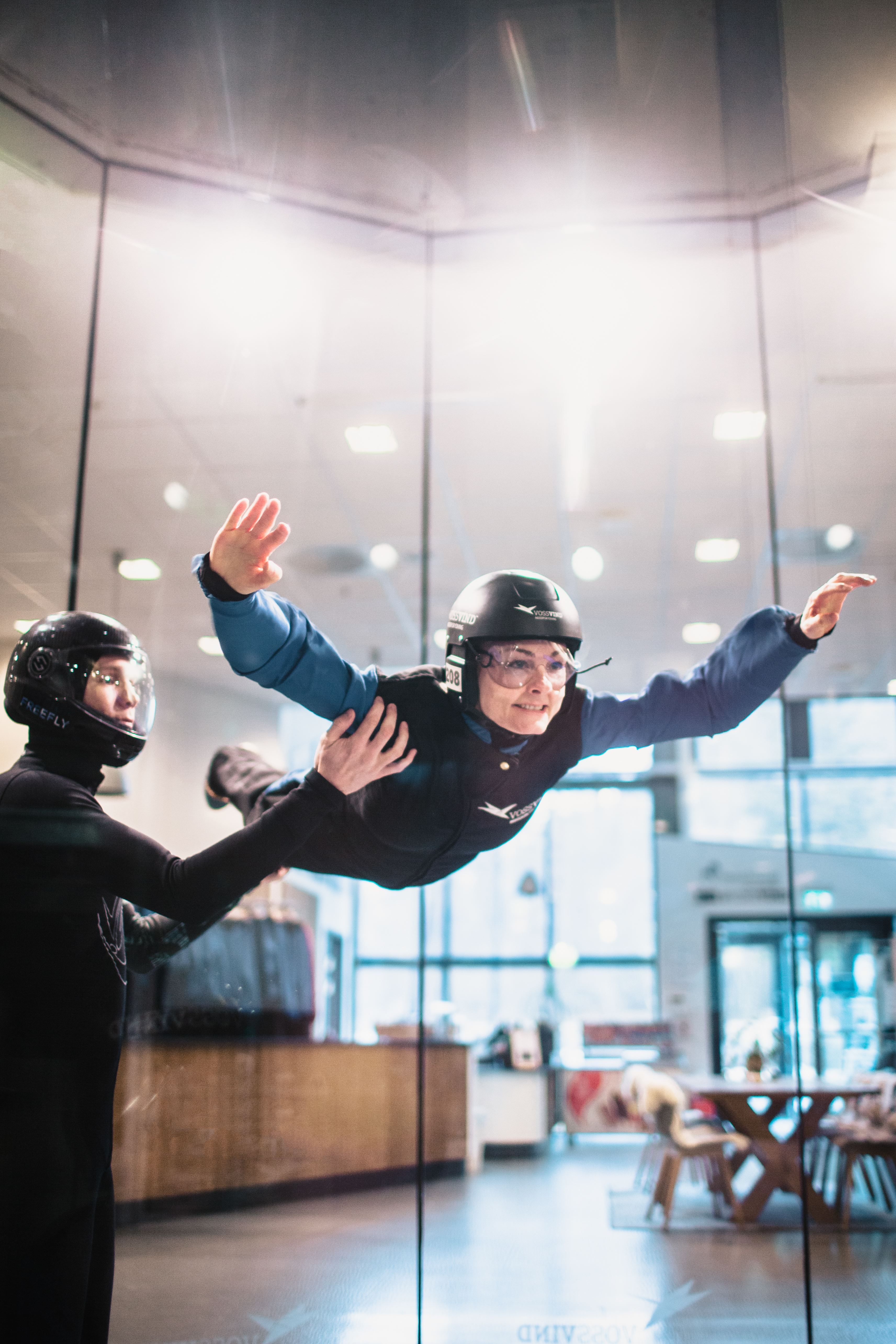 First‑time flyer guided by an instructor inside the glass‑enclosed wind tunnel, arms and legs extended for stability.
