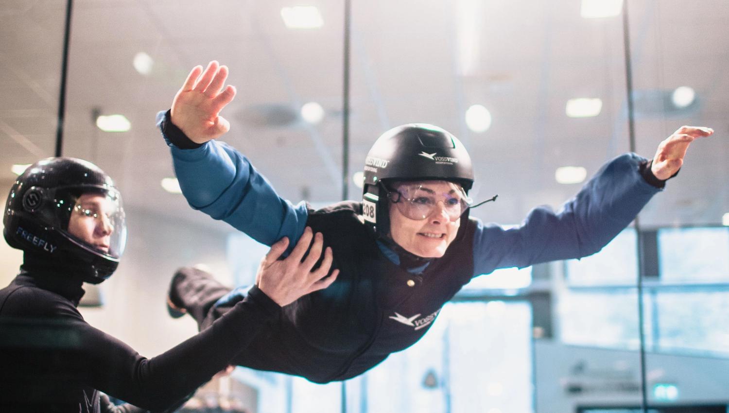First‑time flyer guided by an instructor inside the glass‑enclosed wind tunnel, arms and legs extended for stability.